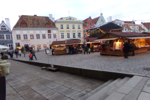 Ein lebendiger Weihnachtsmarkt in Tallinn, Estland, mit Menschen um beleuchtete und geschmückte Stände, Gebäuden mit Fenstern im Hintergrund, Treppen, die zum Markt führen, und Töpfen mit Pflanzen herum.