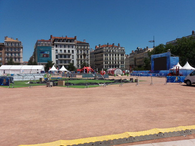 Großer städtischer Platz voller Zelte, umgeben von Gebäuden, Bäumen und Fahrzeugen, unter einem klaren blauen Himmel.
