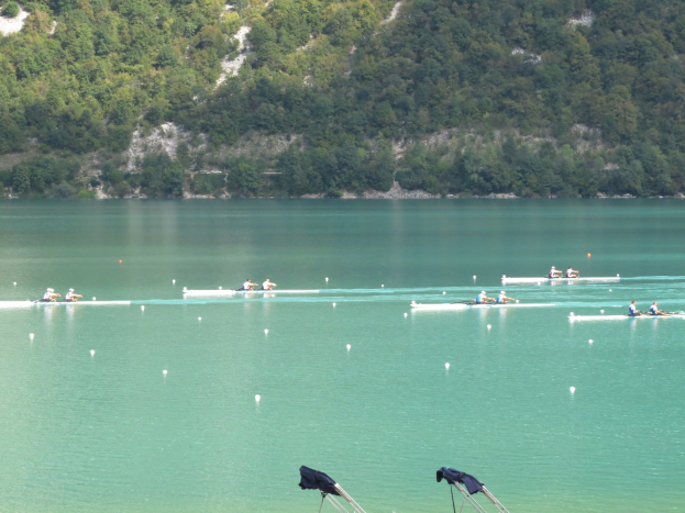 Eine Gruppe von Menschen, die bei einem Wettbewerb auf einem Gewässer mit Bäumen und einem Hügel im Hintergrund rudern, mit Kleidung, die über Stühle am Boden gehängt ist.