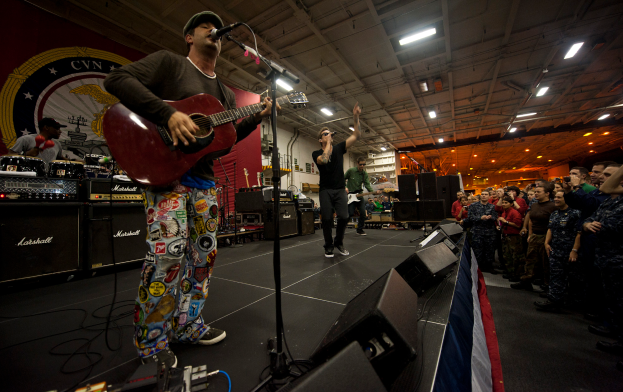 Chris Stapleton, mit einer Mütze bekleidet, spielt Gitarre auf der Bühne vor einem Publikum, umgeben von einem Mikrofon, Lautsprechern, einer Flagge und anderen Instrumenten, mit einem Banner und Lichtern im Hintergrund.
