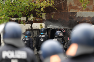 Gruppe von Polizisten in schwarzen Uniformen und Helmen, die vor einem mit Maschendrahtzaun umgebenen Geb├Ąude stehen, mit Bäumen im Hintergrund.