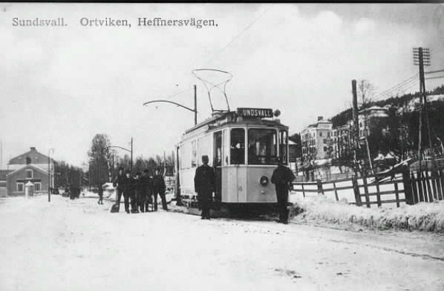Ein Schwarz-Weiß-Foto einer Tram auf einer verschneiten Straße mit Menschen daneben, ein Zaun auf der rechten Seite und Gebäude, Bäume, Pfosten, Drähte und den Himmel im Hintergrund, mit Text oben.
