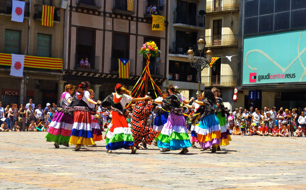 Eine Gruppe Frauen in bunten Kleidern tanzt auf der Straße, umgeben von einer Menschenmenge, mit Gebäuden, Geländern, Balkonen, Fahnen, einem Laternenpfahl und einer Tafel mit Schrift im Hintergrund.