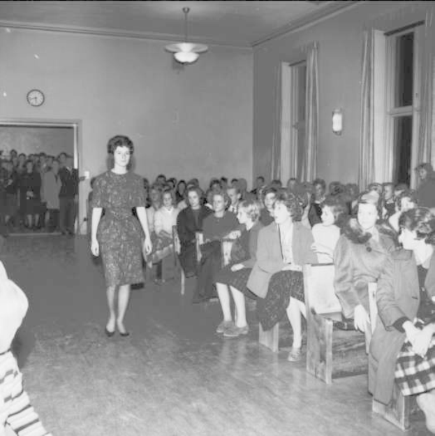 Eine Frau in einem schwarzen und weißen Kleid geht über den Laufsteg während einer Modenschau für Frauenrechte, mit einer Menge, die auf Stühlen sitzt und einer Uhr und Lichtern an den Wänden.