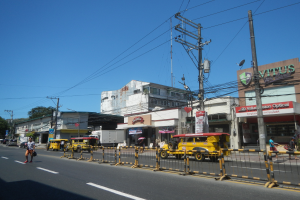 Eine belebte Stadtstraße mit starkem Verkehr, Fußgänger auf dem Gehweg, hohe Gebäude, Bäume und einen klaren blauen Himmel.