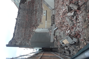 Teilweise abgerissenes Gebäude mit verstreuten Ziegeln und Metallstangen, mit anderen Gebäuden und einem klaren blauen Himmel im Hintergrund.