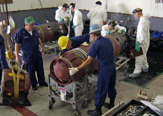 Eine Gruppe von Männern in Helmen und Handschuhen, die eine große Bombe in einer Fabrik mit Maschinen und Wandtext auseinandernimmt.