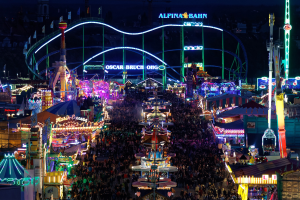 Große Menschenmenge bei nächtlichem Karneval mit bunten Lichtern, Fahrgeschäften, Pfählen, Fahnen und Namensschildern im Dunkeln