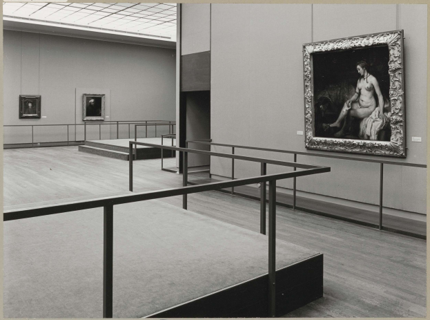 Schwarzes und weißes Foto einer Frau, die in einem Museum voller Rahmenfotos an Wänden, Geländern, Stufen und einem Dach steht, mit Text unten.