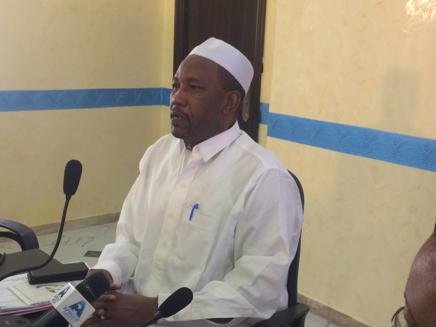 Ein Mann in weißem Anzug und Hut spricht an einem Tisch mit Mikrofonen, Papieren und einer Person mit Brille in einem Raum mit einer Tür im Hintergrund.