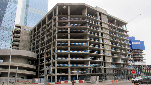 Großes Bauwerk im Bau in einer Stadt umgeben von Wolkenkratzern, mit Fahrzeugen, Fußgängern, Verkehrsinfrastruktur und Kränen gegen einen klaren Himmel.