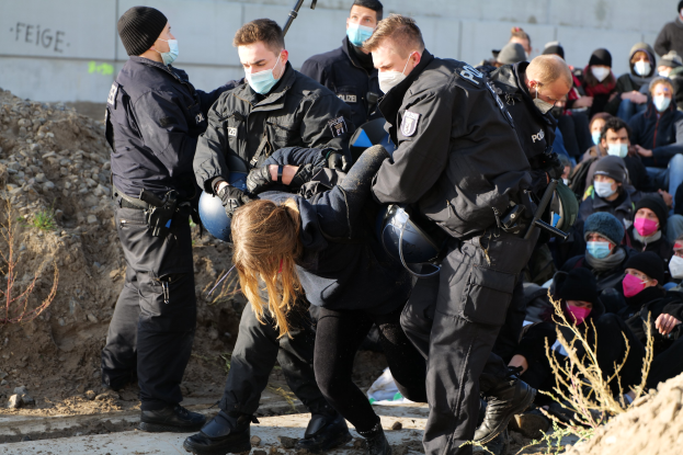 Gruppe maskierter Personen in Kapuzenpullis, einige bewaffnet, die eine Frau umgeben, die von der Polizei eskortiert wird, mit Pflanzen, Steinen und einer Wand mit Protesttext im Hintergrund.