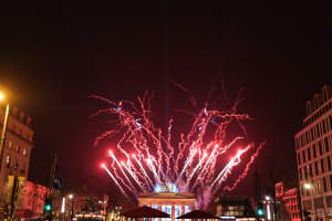 Eine belebte Stadtstraße in Berlin am Neujahrstag, voller Menschen, Fahrzeuge und Gebäude, beleuchtet von Lichtern und Feuerwerk am Himmel.