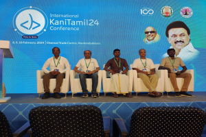 Eine Gruppe von Menschen sitzt vor einem Podium mit Mikrofon, hinter einem Banner mit Text und Bildern von Menschen auf der Internationalen Kanitamil24-Konferenz.