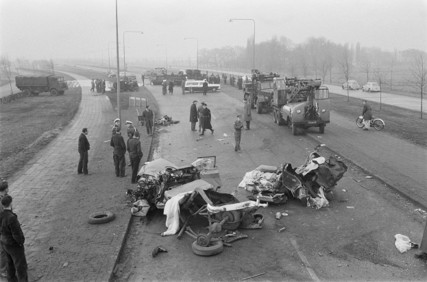 Schwarze und Weiße Szenerie eines Autounfalls am Straßenrand mit mehreren Fahrzeugen, einer Gruppe von Menschen in der Nähe, Laternenpfählen, Bäumen und Himmel im Hintergrund.
