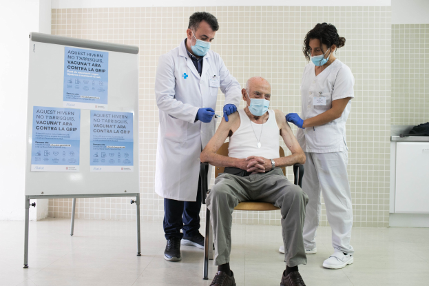 Alter Mann in einem Stuhl sitzend, der von zwei maskierten Ärzten in einem Krankenhauszimmer eine Impfung erhält, mit einer Tafel und Gegenständen auf einer Plattform daneben.