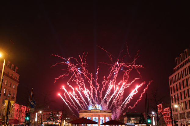 Eine belebte Straßen Szenerie in Berlin an Silvester, voller Menschen, Fahrzeuge und Gebäude, beleuchtet von Feuerwerk und Lichtern, die eine festliche Atmosphäre schaffen.