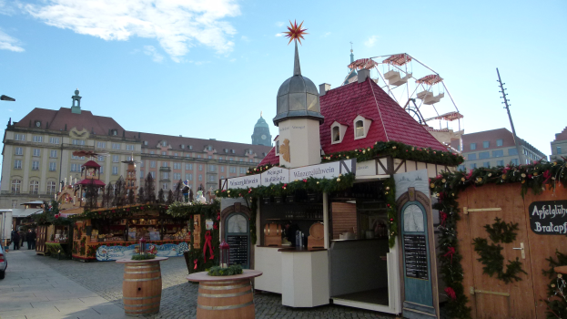 Ein belebter Weihnachtsmarkt auf einem Stadtplatz mit geschmückten Bäumen, Gebäuden, Fahrzeugen, Passanten und bewölktem Himmel.
