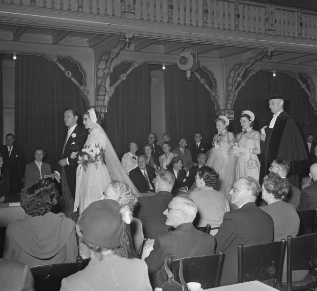 Schwarz-weißes Foto einer Hochzeitszeremonie in einem großen Raum mit sitzenden und stehenden Gästen, einige halten Blumensträuße, Tische mit Tassen und Gegenständen, Vorhänge und Deckenleuchten im Hintergrund.