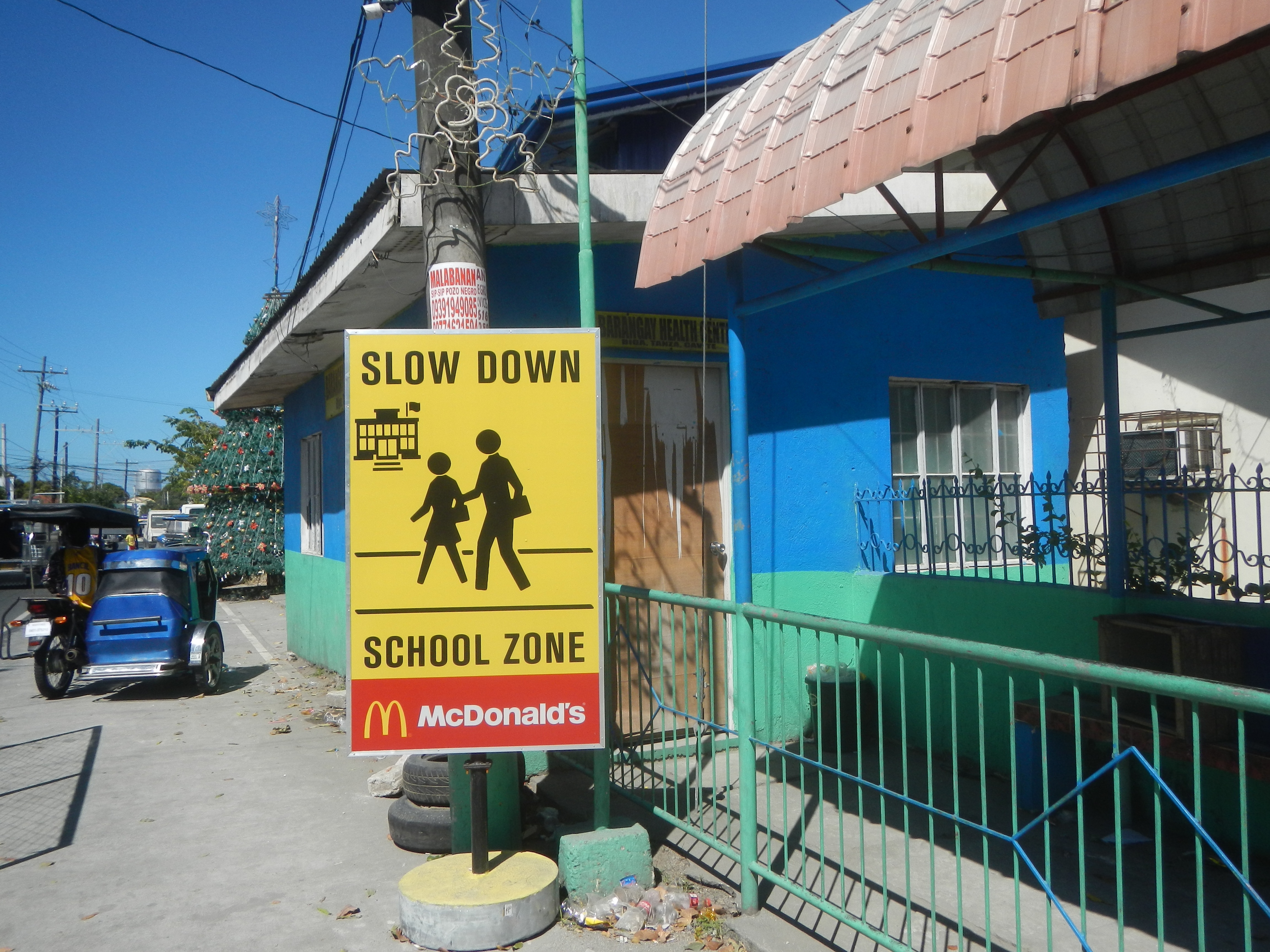 Ein Schulgebäude mit einem "Slow Down"-Schild, Fahrzeuge auf der Straße, ein Geländer, ein Pfahl mit einer Tafel, Bäume, Strommasten mit Drähten und den Himmel im Hintergrund.