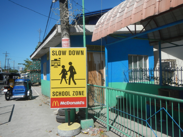 Ein Schulgebäude mit einem "Slow Down"-Schild, Fahrzeuge auf der Straße, ein Geländer, ein Pfahl mit einer Tafel, Bäume, Strommasten mit Drähten und den Himmel im Hintergrund.