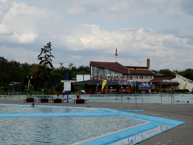 Ein großes Schwimmbad mit Menschen darin, umgeben von Pfosten, Bänken, Topfpflanzen, einem Schild, einem Fahnenmast mit Flagge, einem Gebäude mit Fenstern, Straßenlaternen, einer Baumgruppe und einem bewölkten Himmel.