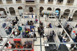 Eine große Gruppe von Menschen versammelt sich in einem Raum, der mit Ständen, Bannern, Tischen, Stühlen und Ballons gefüllt ist, mit einem Gebäude im Hintergrund, das Bögen, Säulen, Geländer und einen Bildschirm aufweist.