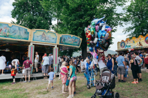 Eine Menschenmenge, darunter einige mit Kinderwagen, umringte einen bunten, beleuchteten Karussell auf einem Volksfest, mit Bäumen und einem bewölkten Himmel im Hintergrund.