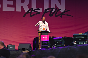 Ein Mann in einem weißen Hemd und braunen Hosen steht auf einer Bühne und hält ein Mikrofon in der Hand, vor ihm eine pinkfarbene Fahne mit der Aufschrift "Berlin as Fuck".