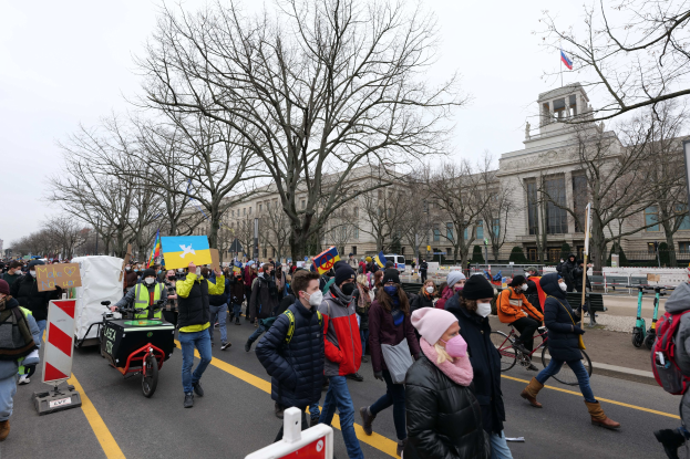 Eine große Gruppe von Menschen marschiert auf einer Stadtstraße, einige halten Schilder und andere fahren Fahrräder, mit Bäumen und einem klaren blauen Himmel im Hintergrund.