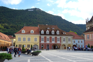 Menschen gehen auf einem Stadtplatz umgeben von Gebäuden mit Fenstern, Laternenmasten und Topfpflanzen, mit einem Hügel und bewölktem Himmel im Hintergrund.