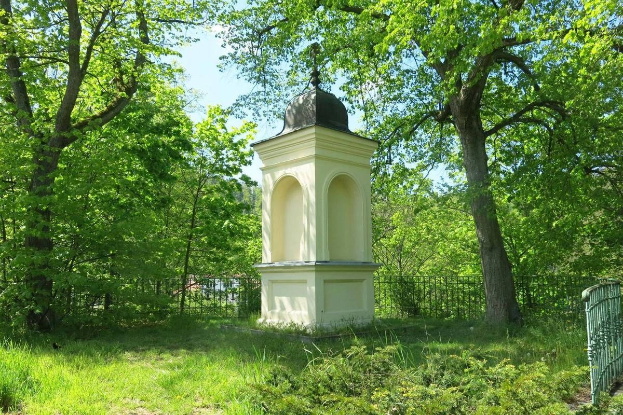 Kleines weißes Holocaust-Gedenkmal auf einem grasbewachsenen Friedhof, umgeben von einem Zaun und Bäumen, unter einem klaren blauen Himmel in Vilnius, Litauen.