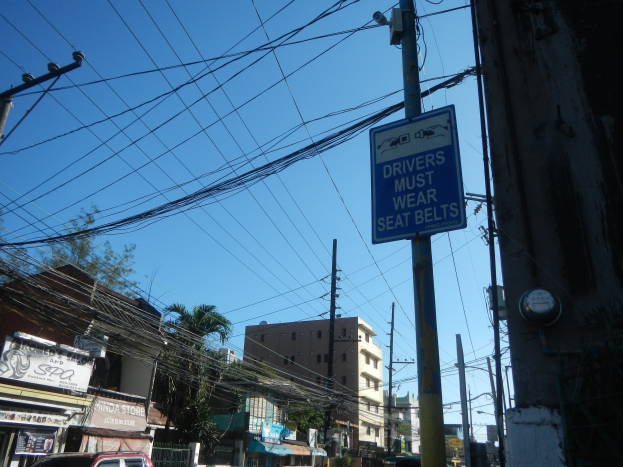 Stadtstraße mit fahrenden Autos, Strommäste mit Drähten, Gebäude, Bäume und Namensschilder, mit einem "Fahrer müssen den Gurt anlegen"-Schild an einem Strommast.