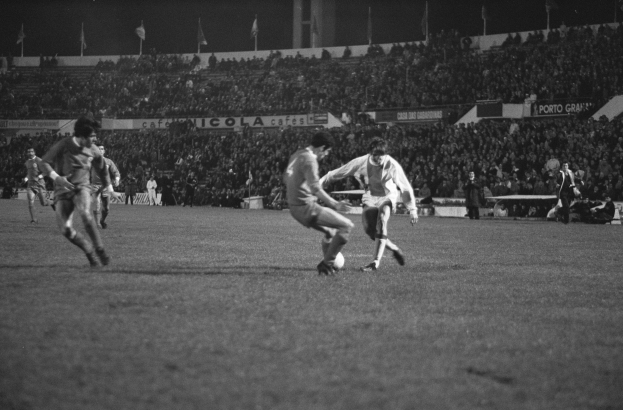 Schwarzes und weißes Bild von Männern, die Fußball in einem Stadion mit Zuschauern auf den Rängen und Fahnen im Hintergrund spielen.