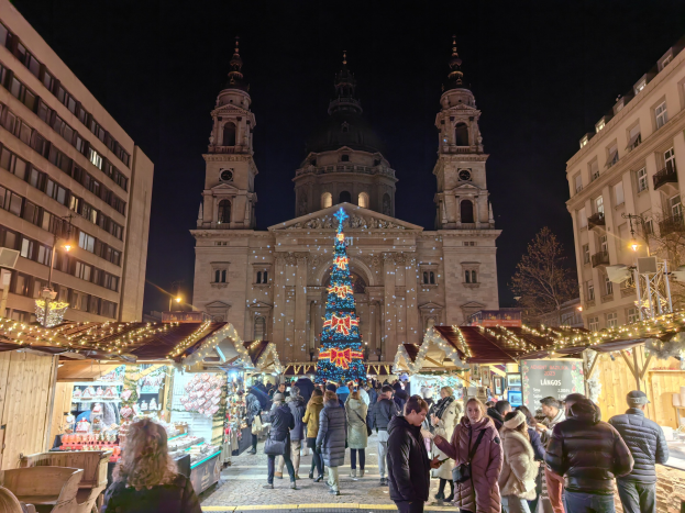 Ein belebter Weihnachtsmarkt mit festlich geschmückten Ständen, die vor einer Kirche in der Nacht beleuchtet sind, umgeben von Gebäuden, Bäumen und einem sternenklaren Himmel.