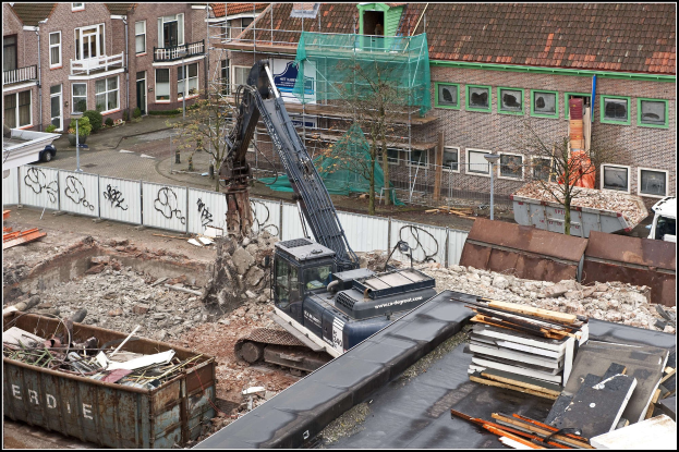 Bagger in Betrieb auf einer Baustelle umgeben von Gebäuden, Grünflächen, Infrastruktur und Fahrzeugen, mit Text unten.
