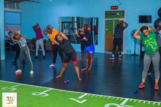 Eine Gruppe von Menschen, die Yoga in einem Fitnessstudio macht, steht auf dem Boden mit Hanteln, umgeben von Sportgeräten, einem Fernseher, einem Spiegel und Deckenleuchten, mit einem Logo und Text auf der linken Seite.