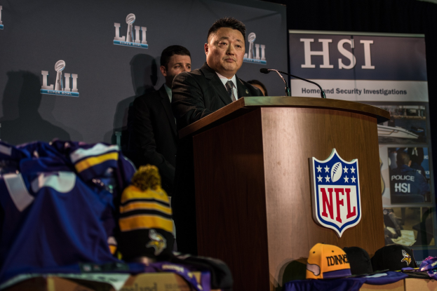 Ein Mann in einem schwarzen Blazer, wei├čem Hemd und schwarzer Krawatte spricht an einem Pult mit Mikrofon und Logo vor einer Menge; blaue und gelbe Merchandise und eine "Super Bowl LII"-Aufschrift sind im Hintergrund zu sehen.
