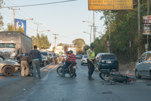 Eine Gruppe von Menschen steht um ein verunglücktes Motorrad auf der Seite einer Straße mit mehreren Fahrzeugen, darunter ein Lastwagen, und einem Hintergrund aus Bäumen, Polen, Lampen und Schildern unter dem Himmel.