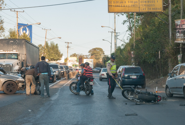 Eine Gruppe von Menschen steht um ein verunglücktes Motorrad auf der Seite einer Straße mit mehreren Fahrzeugen, darunter ein Lastwagen, und einem Hintergrund aus Bäumen, Polen, Lampen und Schildern unter dem Himmel.