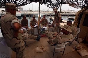 Gruppe von Männern mit Mützen und Schwimmbrillen sitzt um einen Tisch auf einem Boot mit Taschen und einem Zelt in der Nähe, Boote und Gebäude sind im Hintergrund unter einem klaren blauen Himmel zu sehen.