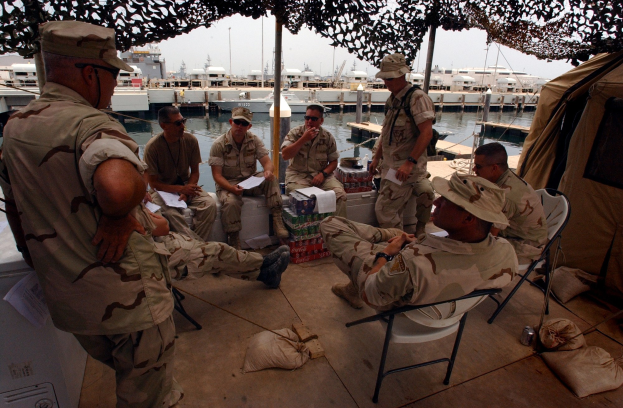 Gruppe von Männern mit Mützen und Schwimmbrillen sitzt um einen Tisch auf einem Boot mit Taschen und einem Zelt in der Nähe, Boote und Gebäude sind im Hintergrund unter einem klaren blauen Himmel zu sehen.