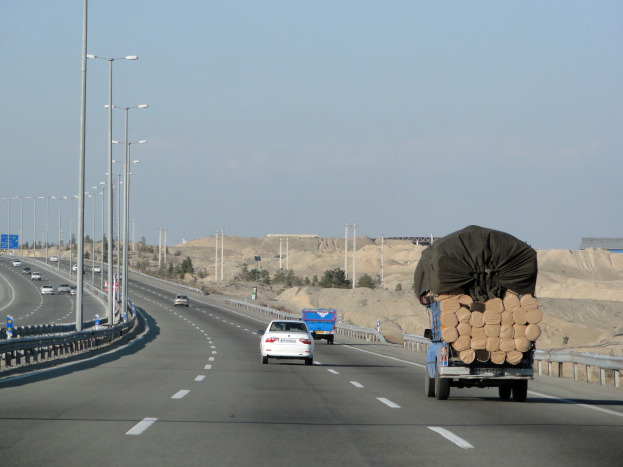 Ein Holztransporter mit einer großen Ladung Holz fährt auf einer Autobahn mit Leitplanken, Laternen, Schildern, Bäumen und Sand, mit Hügeln und einem klaren blauen Himmel im Hintergrund.