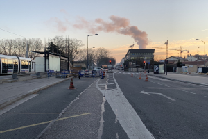 Eine städtische Straße mit Verkehrskegeln, einem Bus, Laternenmasten, Bäumen, Gebäuden, Kränen und Rauch, der aus einem fernen Gebäude aufsteigt.