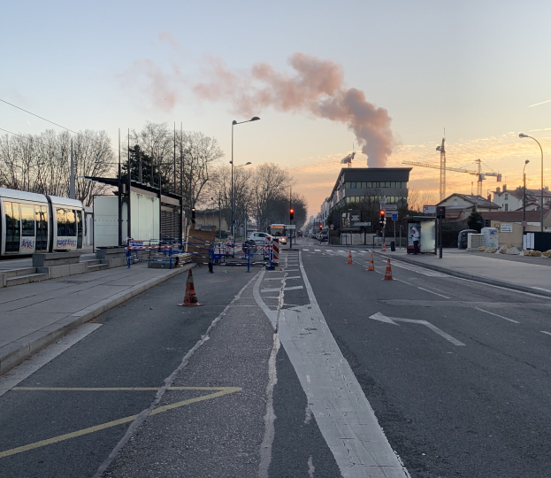 Eine städtische Straße mit Verkehrskegeln, einem Bus, Laternenmasten, Bäumen, Gebäuden, Kränen und Rauch, der aus einem fernen Gebäude aufsteigt.