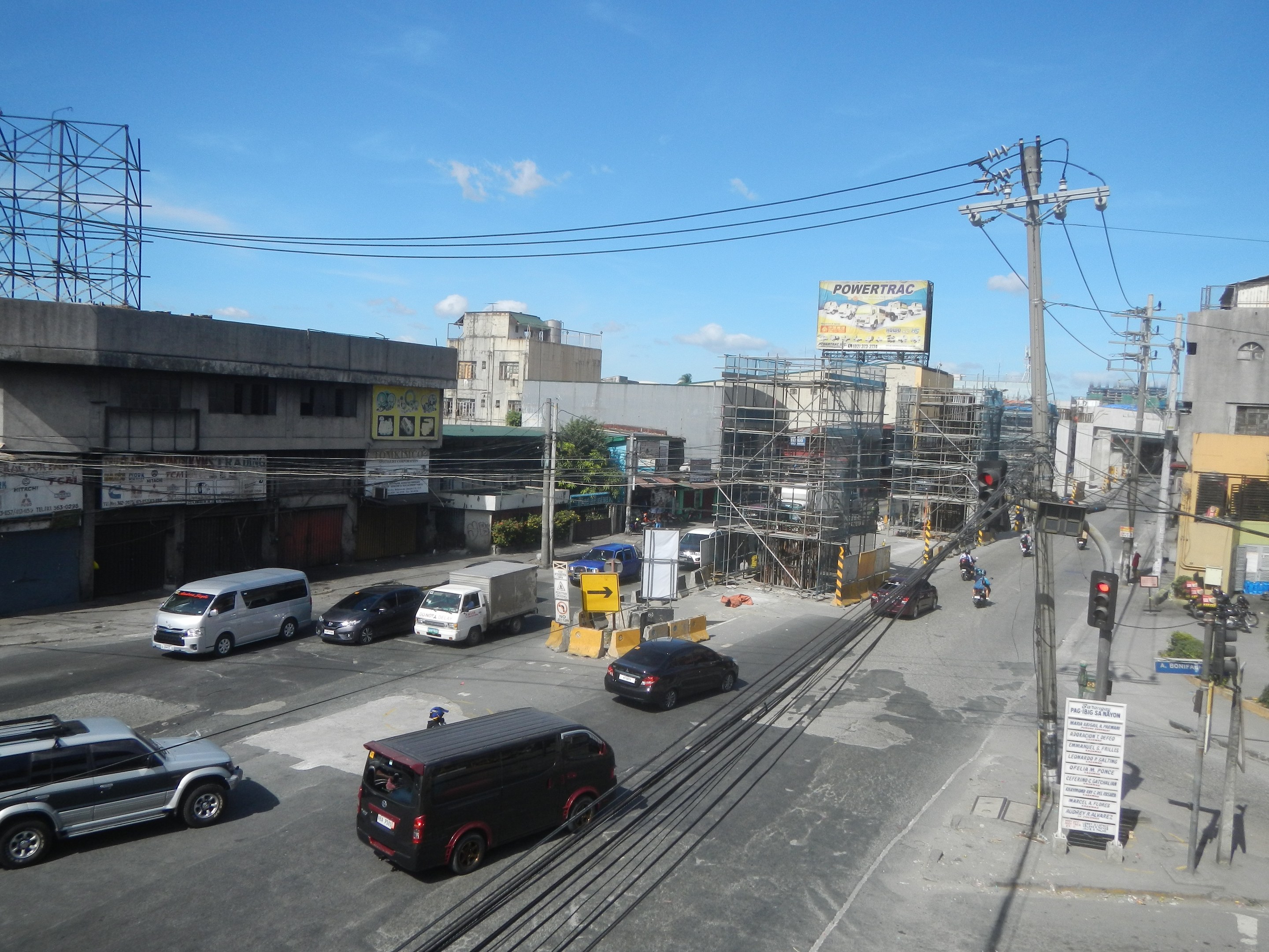 Eine belebte Stadtstraße mit hohen Gebäuden, Fahrzeugen, Strommasten mit Drähten, Verkehrsampeln, Schildern, Bäumen und einem bewölkten Himmel.