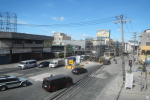 Eine belebte Stadtstraße mit hohen Gebäuden, Fahrzeugen, Strommasten mit Drähten, Verkehrsampeln, Schildern, Bäumen und einem bewölkten Himmel.