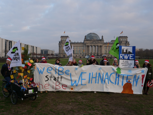 Gruppe von Menschen mit Mützen, die ein Transparent vor dem Reichstaggebäude halten, mit einer Person auf einem Kinderwagen, Bäumen, Gebäuden und Flaggen im Hintergrund.