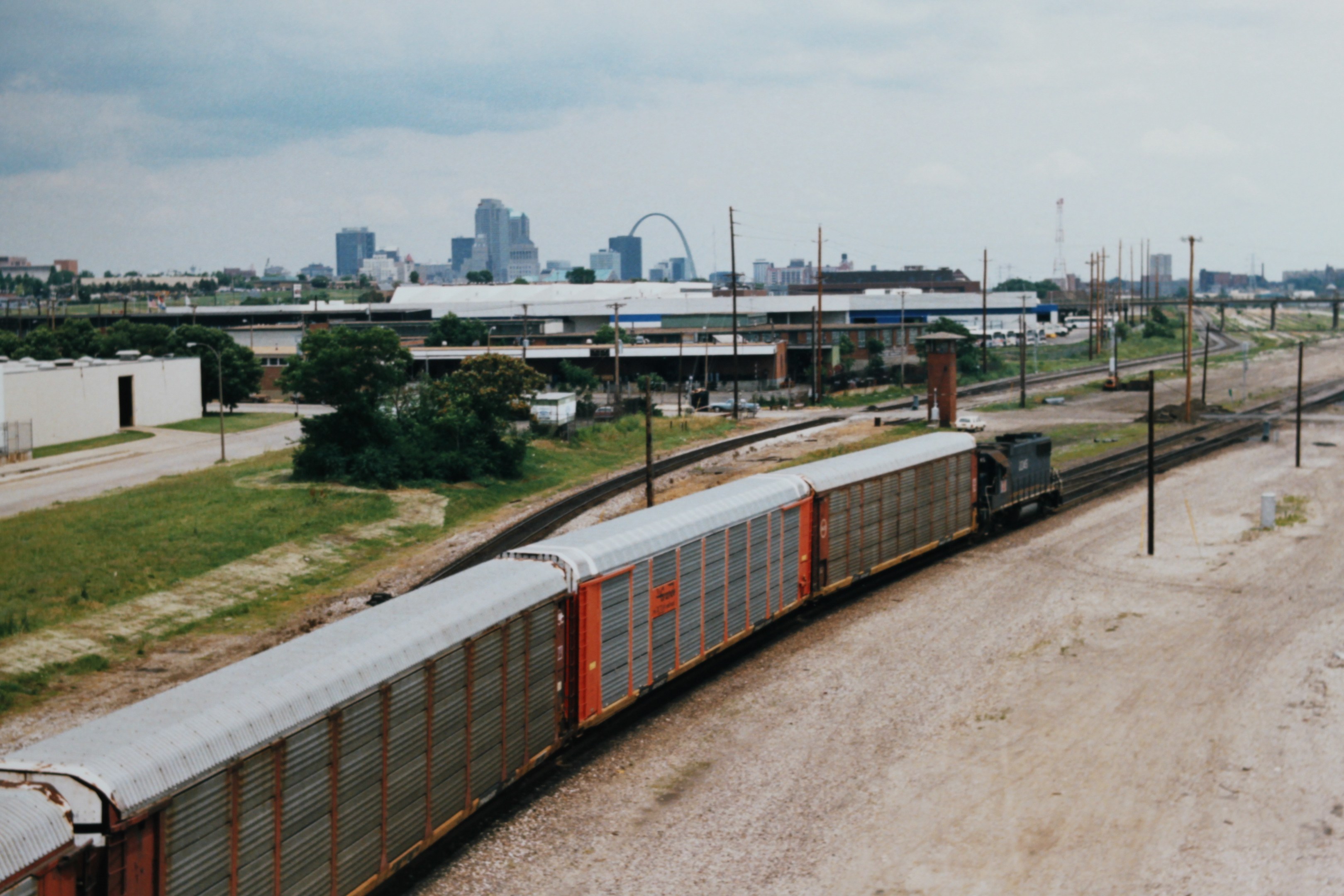 Güterzug fährt auf Schienen neben einer Stadtlandschaft mit Strommasten, Gebäuden, Bäumen, Gras und einem bewölkten Himmel.