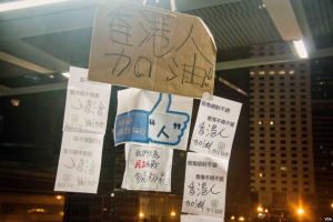 Eine Gruppe von Menschen vor einem Gebäude bei Nacht, beleuchtet von Stadtlichtern, mit mehreren Protestschildern an der Decke.